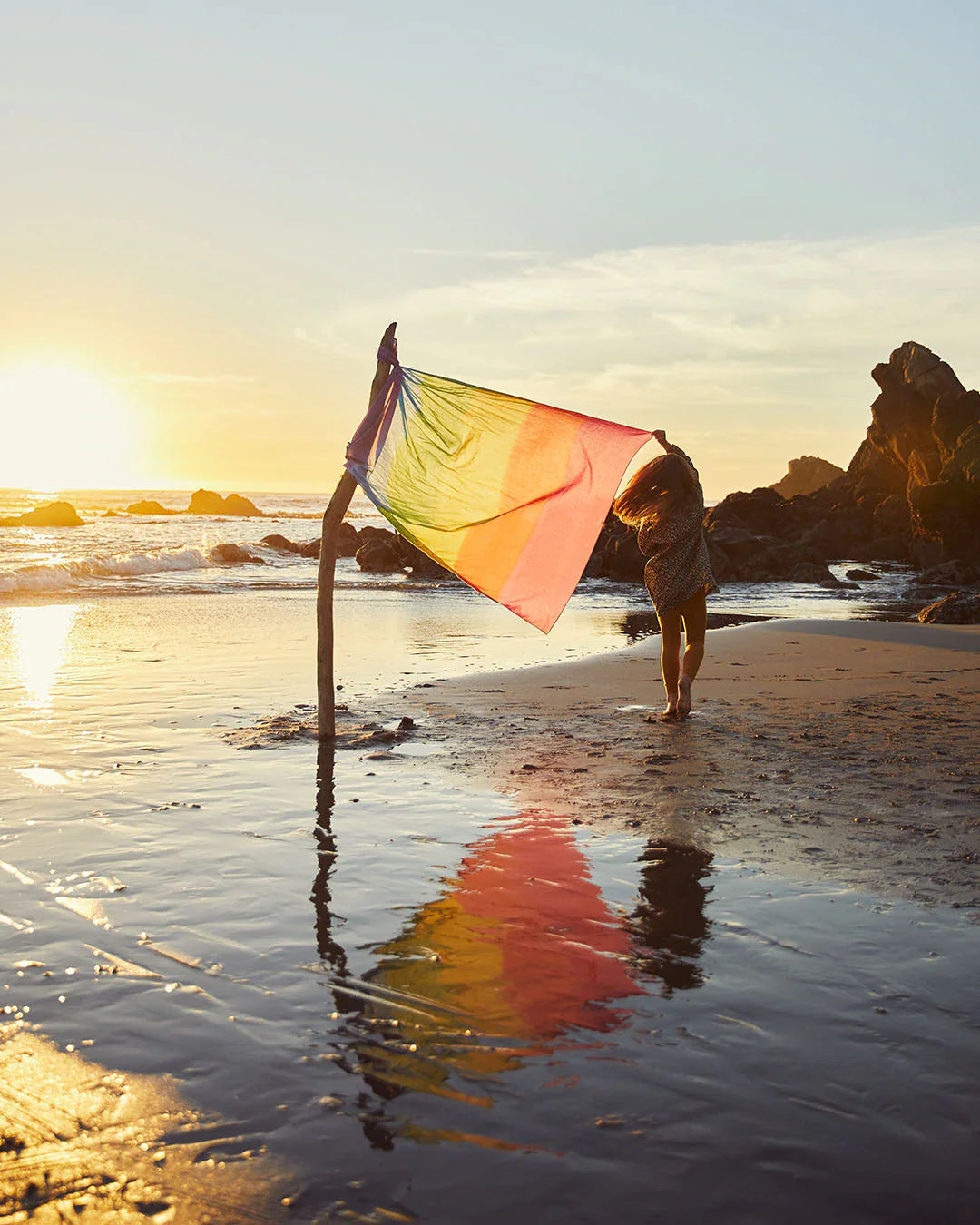 Childs at the beach with the sun setting in the back ground there is a large rainbow play silk in on a branch to create a flag pole standing in the sand