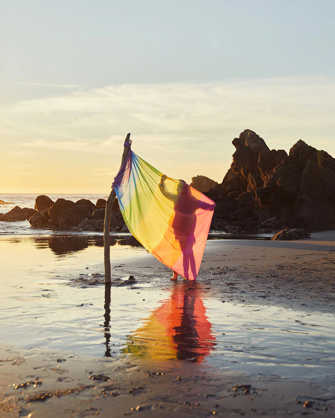 Childs at the beach with the sun setting and rocks in the back ground. The child stand s silhouetted behind a large rainbow play silk in on a branch to create a flag pole standing in the sand