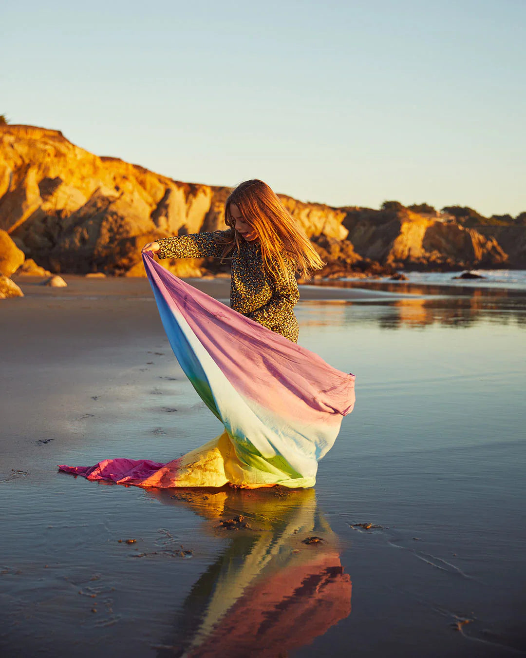 Child planing int he wet sand with a rock out cropping in the back ground. They are spinning with a rainbow cloth play silk that billows and trails int he sand