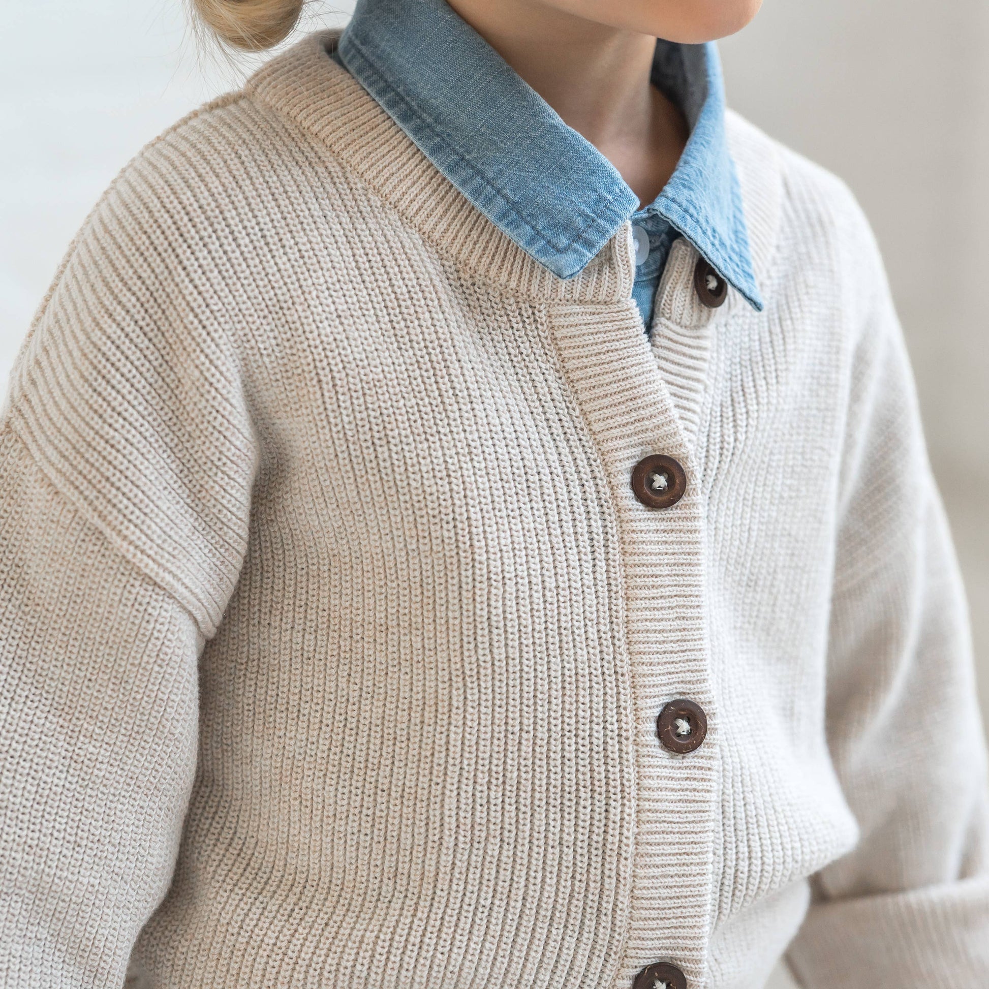 neck detail of child wearing a heather oat knit cardigan buttoned up over a denim shirt-dress