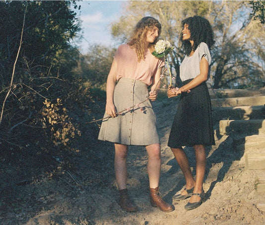 Two femme-presenting people standing outside holding flowers wearing bae skirts by field days and light-colored blouses