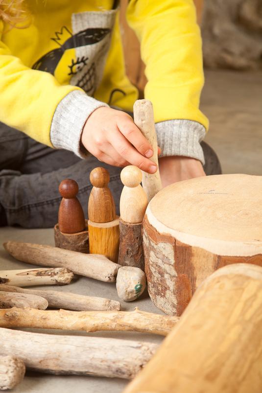 A child sitting on the floor, playing with three wooden peg people, wooden sticks and trunks