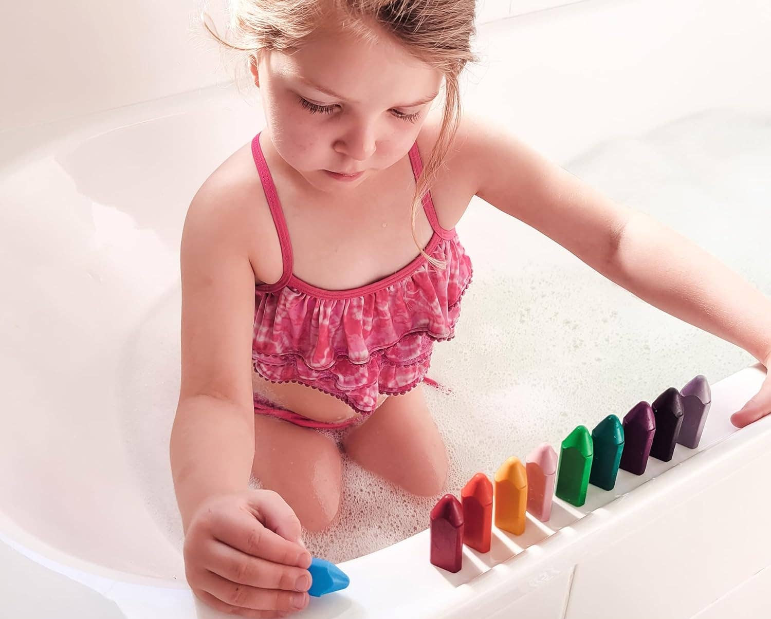 a child wearing a swimsuit taking a bath lining up their triangle bath crayons on the side of the bathtub