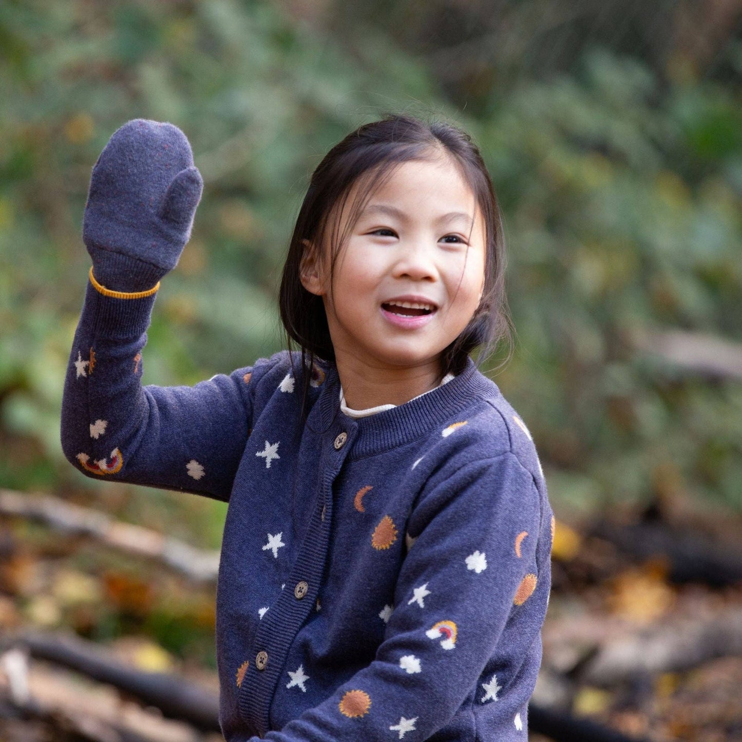 Child sitting outdoors wearing an around the sun organic knitted cardigan and waving