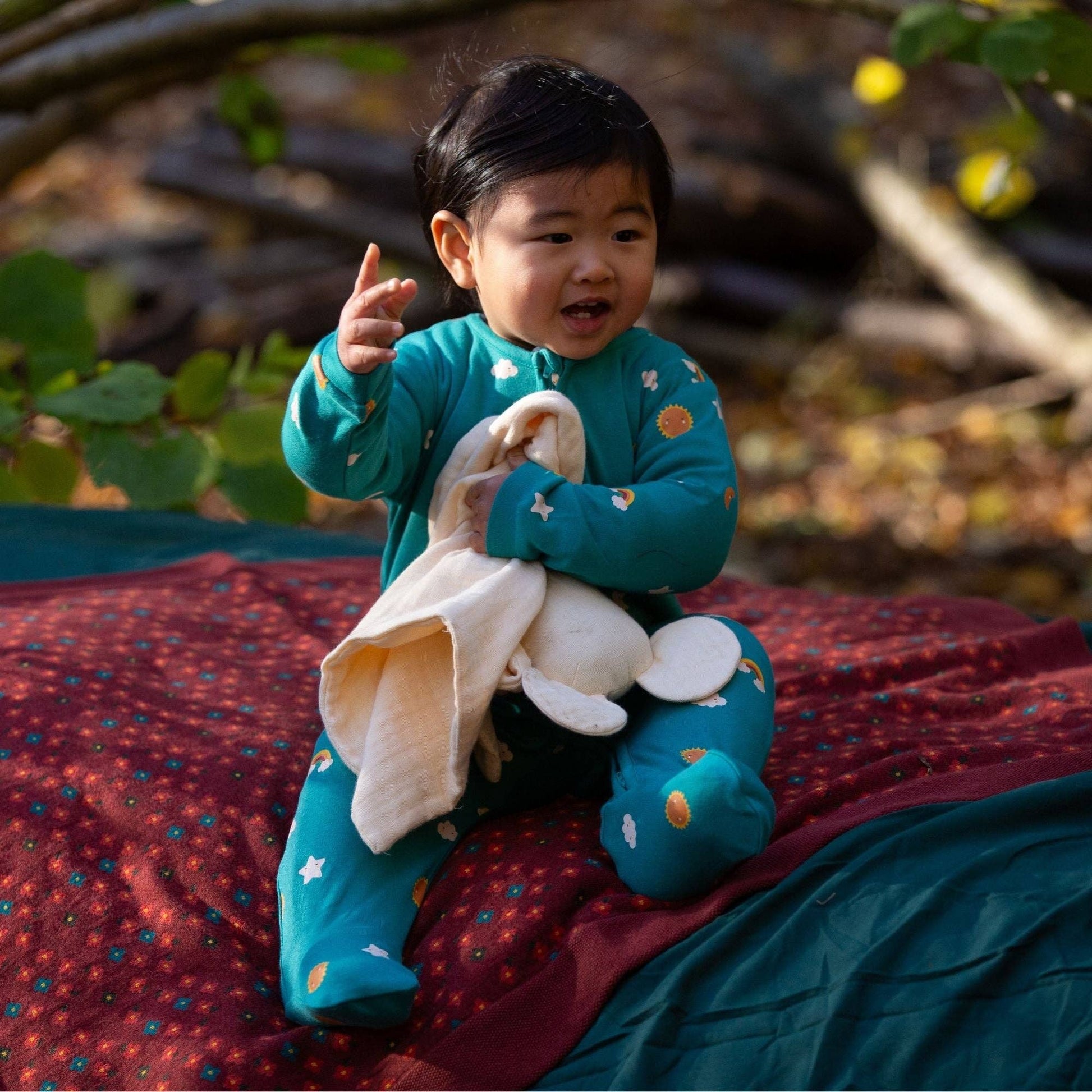 baby sitting outside on some blankets wearing turquoise skies organic footie and holding a lovey