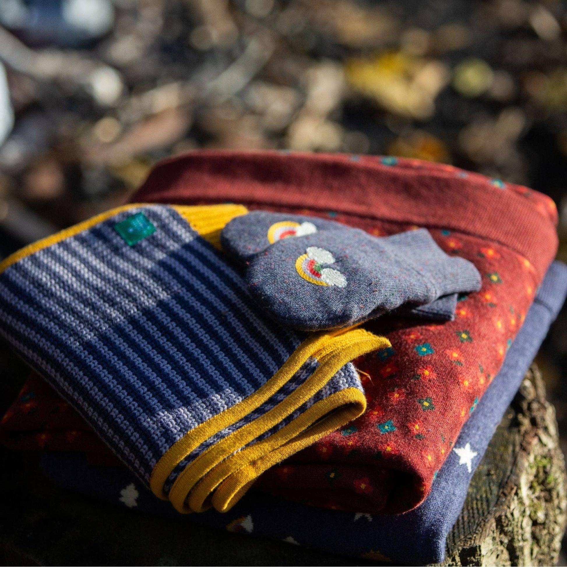 two baby blankets, a scarf, and blue knitted mittens stacked on a log outside