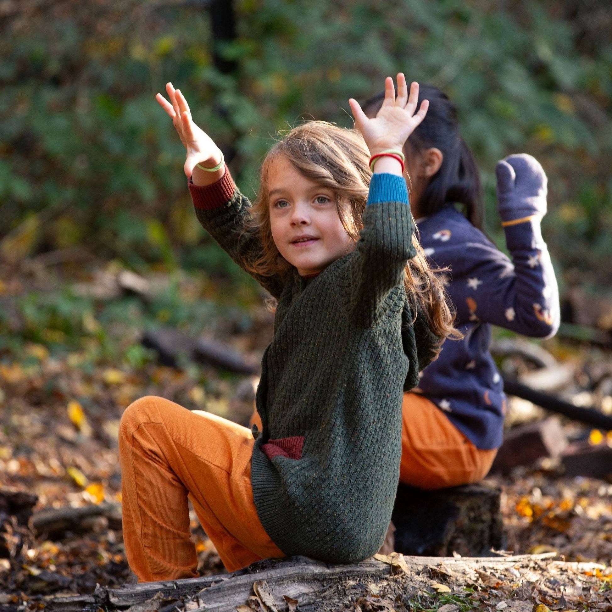 Child sitting outdoors on a tree trunk with their hands in the air wearing a green hooded cardigan