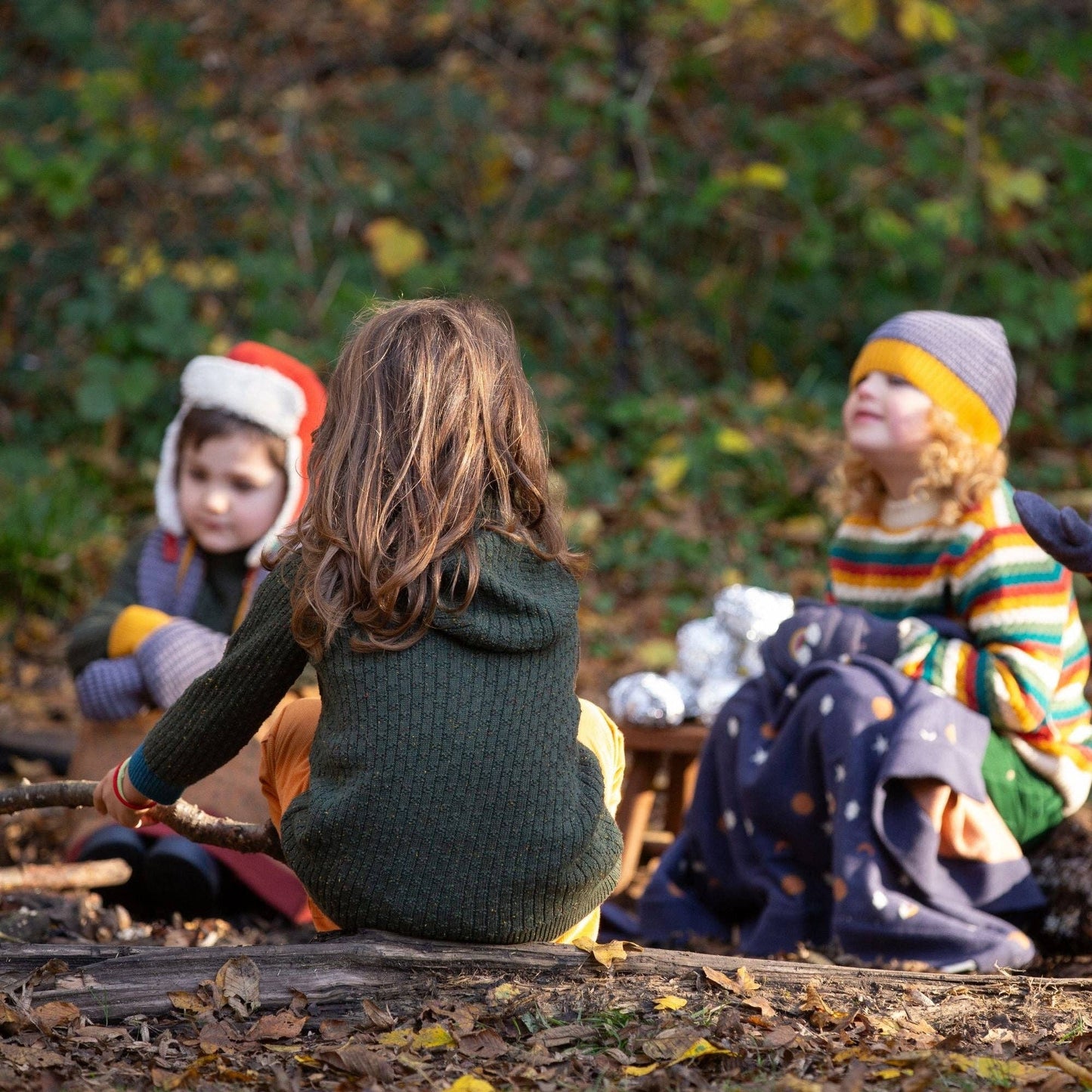 Children sitting outdoors around a fire wearing organic cotton hats and sweaters by little green radicals