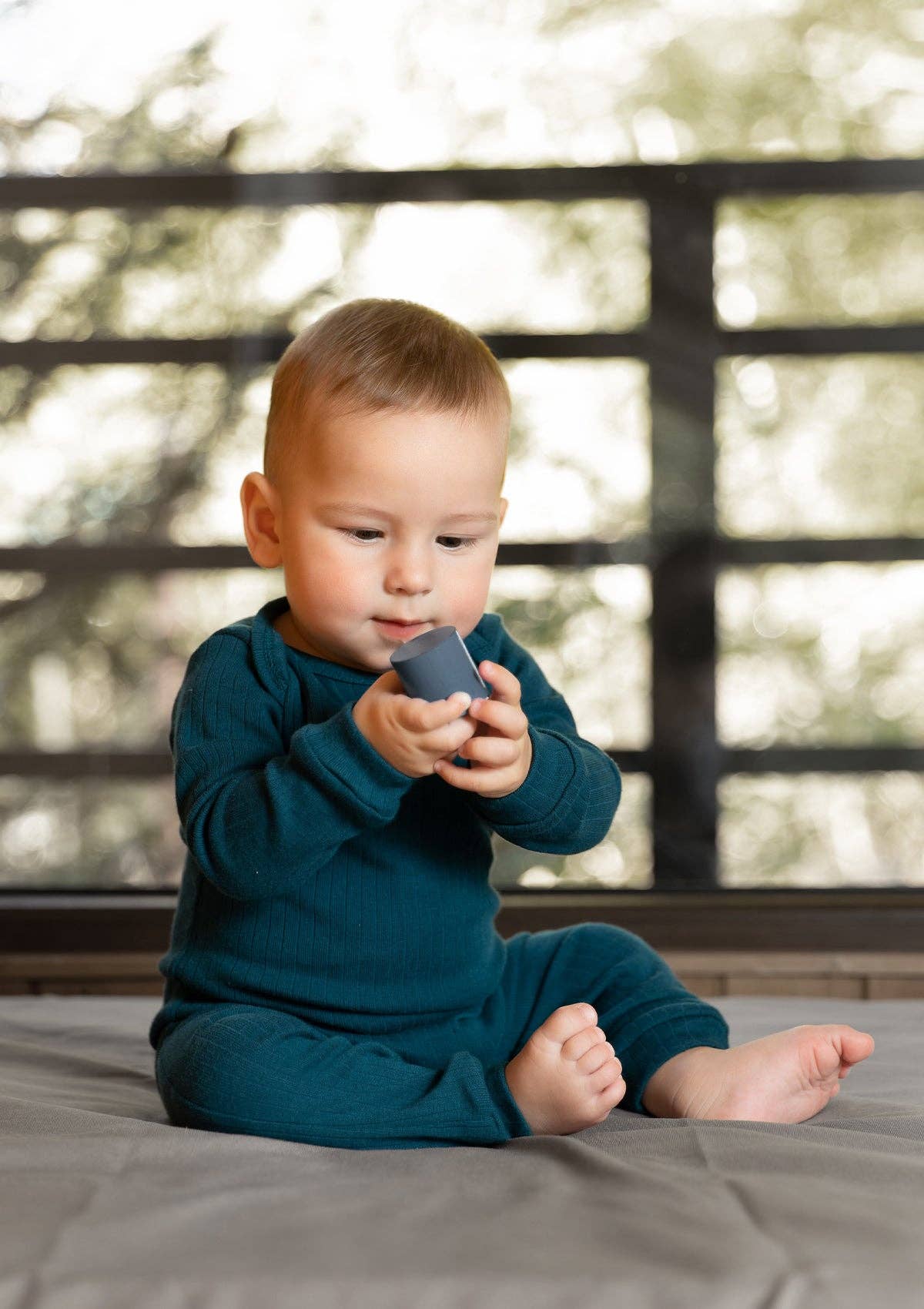 Baby sitting on the floor holding a wooden toy wearing a long sleeve ribbed romper in petroleum blue