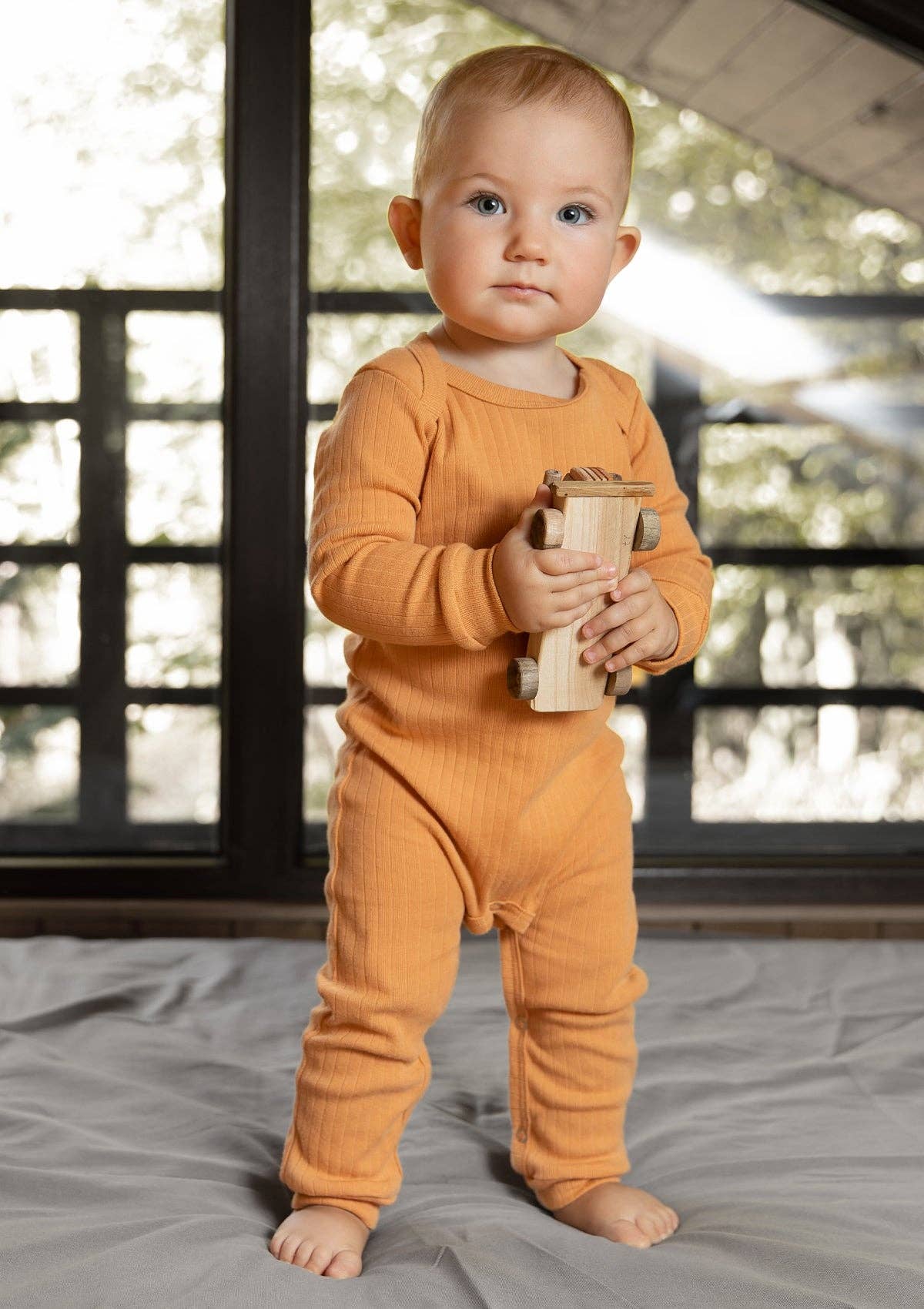 A baby standing on a mat holding a wooden car wearing a long sleeve romper in ochre