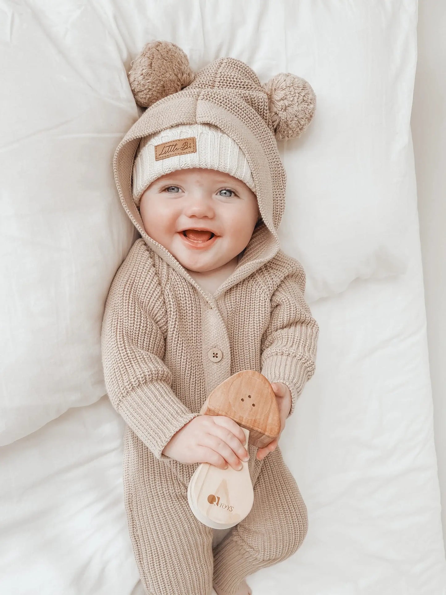 a baby laying on a bed wearing a hooded romper with bear ears holding a wooden mushroom rattle