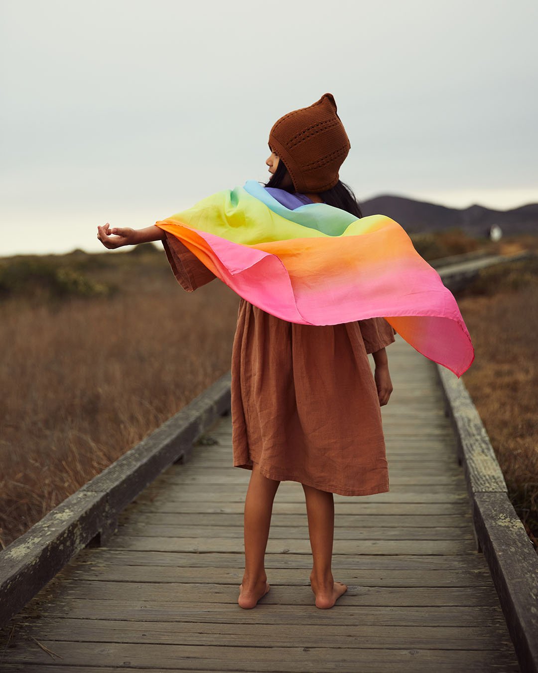 child walking on a boardwalk wearing rainbow playsilk as a cape