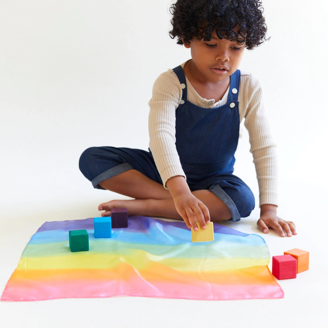 Child sitting on the floor playing with mini rainbow playsilk and wooden blocks